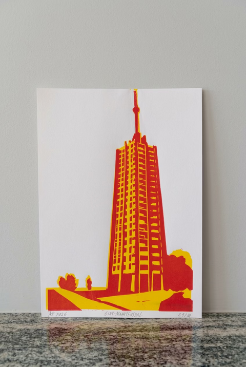 A white art print featuring a stylized red and yellow illustration of the Empire State Building, leaning against a red-brown brick wall on a dark stone surface.