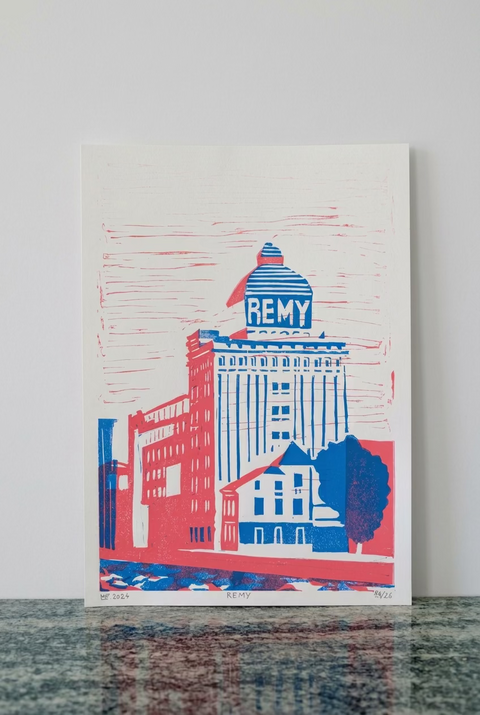 A vertical art print featuring a red and blue architectural illustration of the Remy building, displayed against a red-brown brick wall on a dark stone surface.