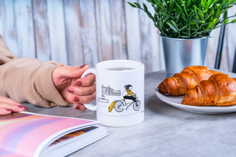 White ceramic mug featuring an illustrated woman riding a bike with a suitcase past a Leuven landmark, styled in black and yellow.