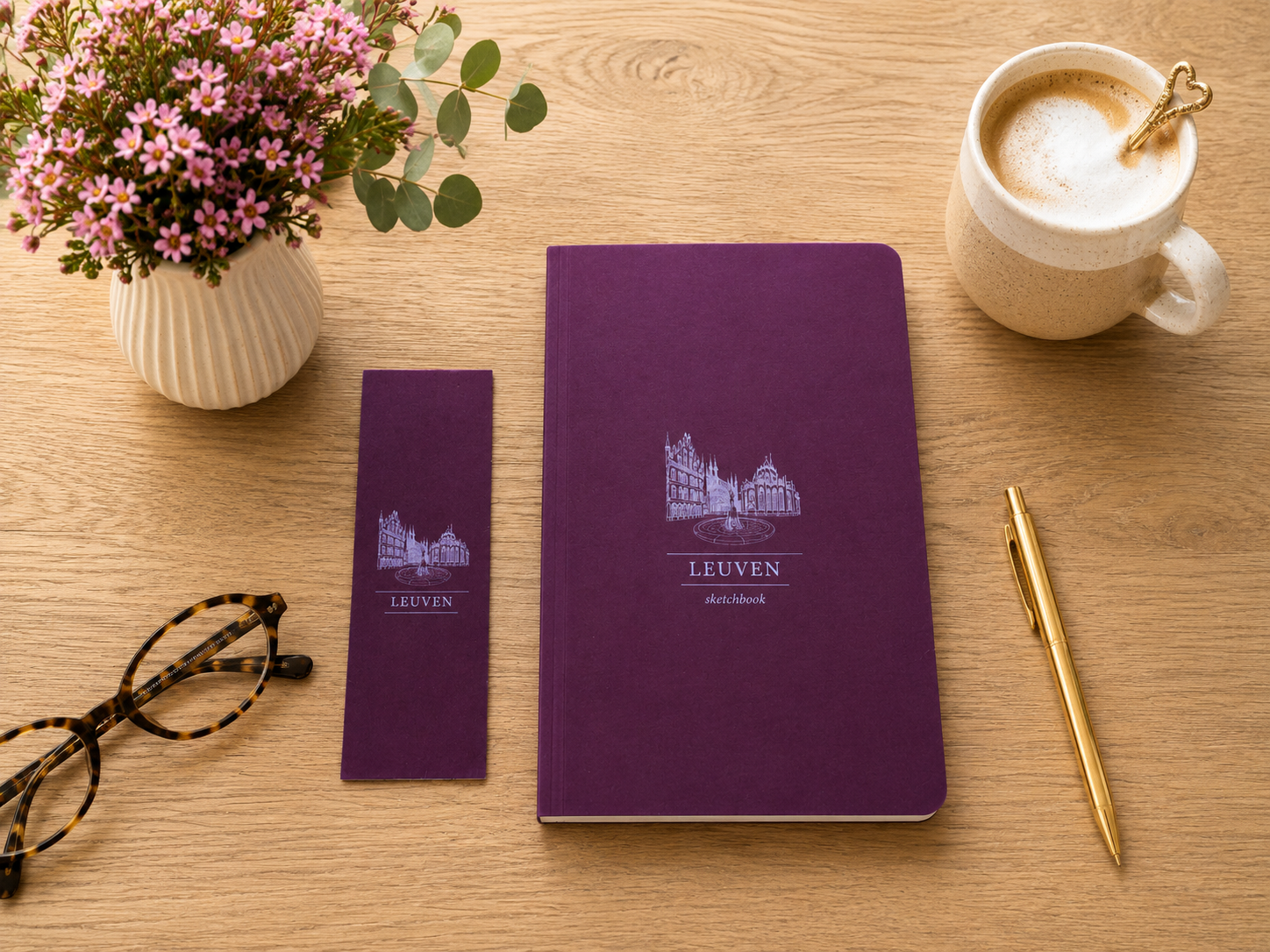 Premium Leuven sketchbook with a matching illustrated bookmark displayed on a warm oak desk. A handcrafted ceramic mug, elegant reading glasses and colourful flowers create a cosy workspace inspired by slow mornings and creative journaling.