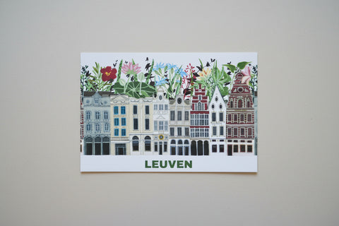 A rectangular art print featuring a colorful illustration of Leuven canal houses with floral details above, mounted on a light beige wall. The word 'LEUVEN' is printed in green at the bottom.