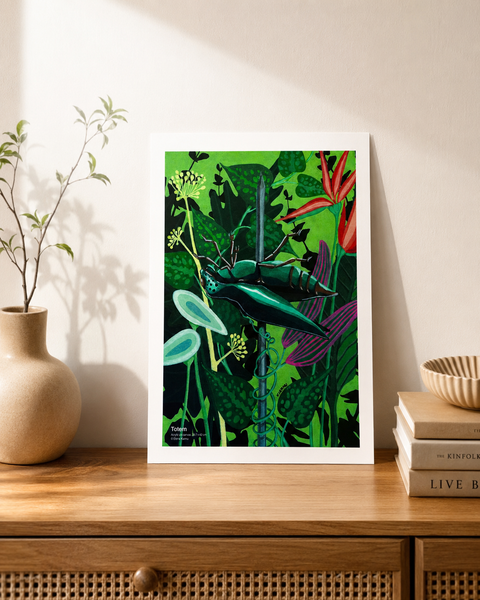 A framed botanical art print featuring a green beetle and tropical leaves, styled on a wooden console table next to a vase and books against a beige wall.