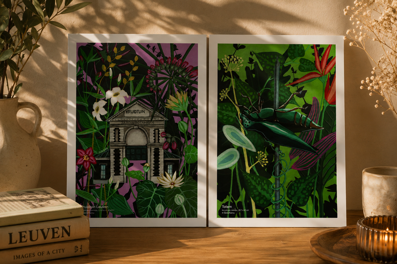 Two framed botanical art prints displayed side by side on a wooden table, surrounded by decorative props, including books, a ceramic vase, and a candle. The left print features a building surrounded by flowers on a purple background, and the right print features a green dragonfly among lush tropical leaves.