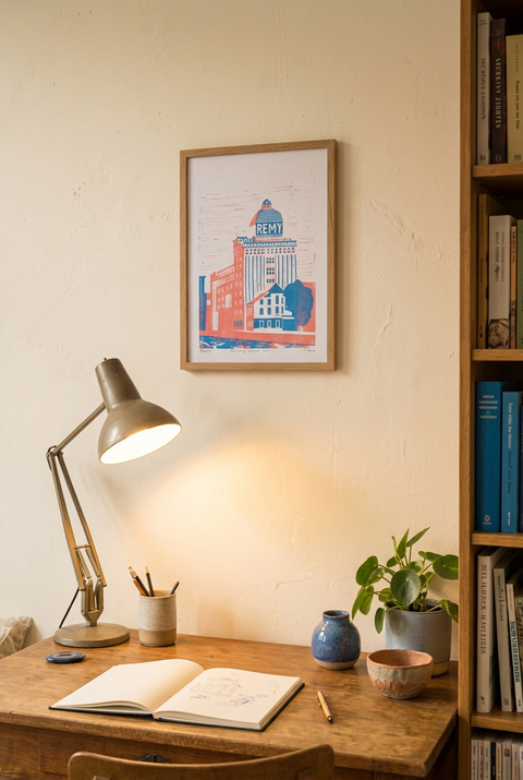 A framed art print featuring a stylized red and blue building illustration with the text 'REMY' hanging on a beige wall above a wooden desk with a lit lamp, open book, and small plants.